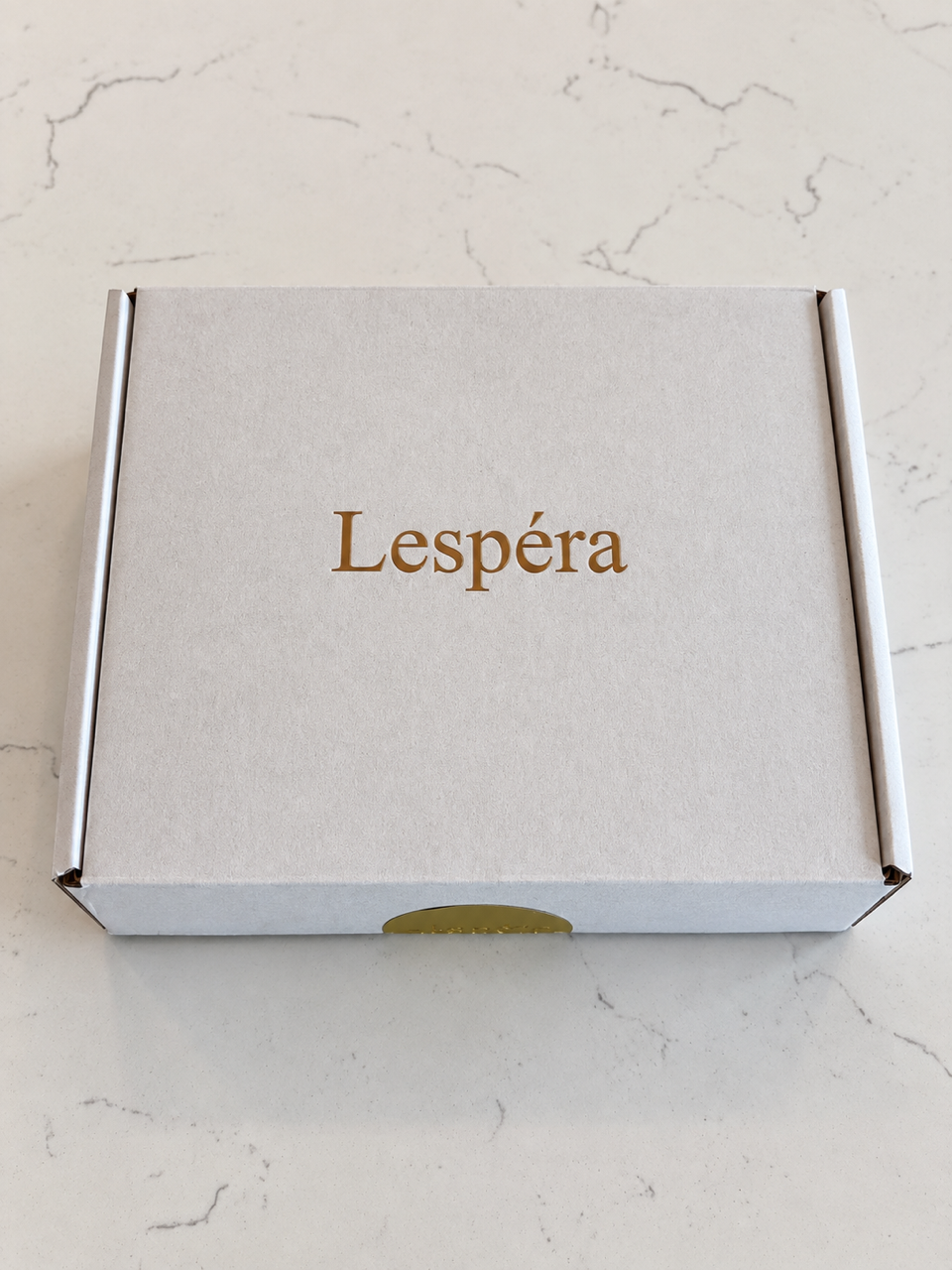 L'écrin cadeau Lespéra fermé, avec le logo Lespéra en or sur une surface blanche douce