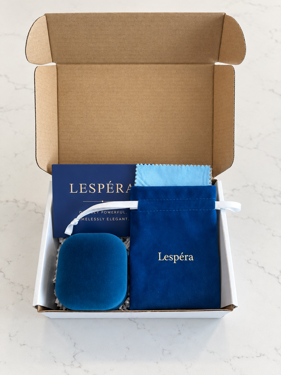 Un écrin cadeau Lespéra ouvert montrant un coffret en velours bleu marine plus petit, une pochette à cordon, un chiffon de polissage et une carte d'entretien